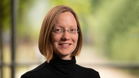Das Foto zeigt das Portrait einer Frau mit schulterlangem blonden Haar und Brille. Sie trägt einen schwaren Rollkragenpullover und lächelt in die Kamera. Der Hintergrund ist verschwommen.