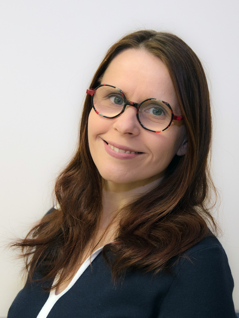 Das Foto zeigt das Portrait einer Frau mit langem braunen Haar und einer Brille mit buntem Gestell. Sie steht vor einem weißen Hintergrund und lächelt in die Kamera.