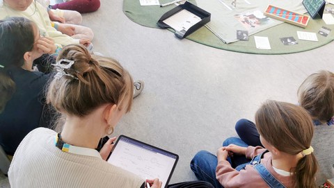 Das Foto zeigt eine Lernsituation, bei der eine erwachsene Person und Kinder auf dem Boden sitzen und von verschiedenen Lernmaterialien umgeben sind.