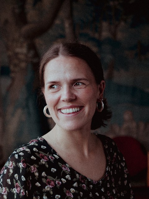 Das Foto zeigt eine Frau, die etwas außerhalb des Bildausschnittes anschaut und freundlich lächelt. Sie hat kürze Haare, auffällige goldene Ohrringe und ein gemustertes Oberteil.