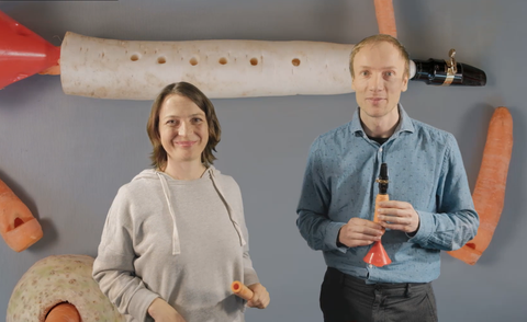 Das Foto zeigt zwei Personen, die freundlich in die Kamera lächeln. Links steht eine Frau, rechts ein Mann, der ein selbstgebasteltes Blasinstrument aus einer Karotte in der Hand hält. Im Hintergrund sind verschiedene Gemüse zu sehen.