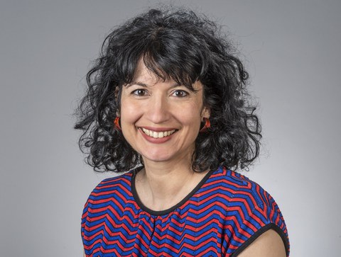 Das Portraitfoto zeigt eine Frau mit schwarzem, gelocktem Haar. Sie trägt rote Ohrringe und ein rot-blau gestreiftes T-Shirt.