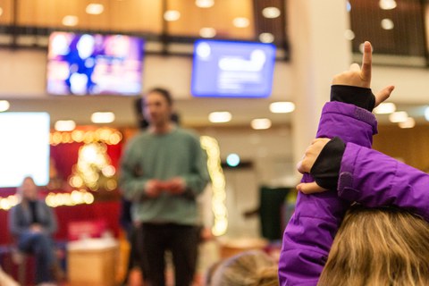 Das Foto zeigt im Vordergrund eine Kinderarm mit einer sich meldenden Hand. Im Hintergrund ist eine Person auf einer Bühne, Bildschirme und Adventsdekoration.