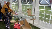 Das Foto zeigt eine Wippe aus Glas und Holz. 2 Kinder stehen davor, eine erwachsene Person hält den einen Sitz der Wippe. Die Wippe steht in einem Festzelt.