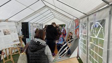 Das Foto zeigt das Innere eines Festzelts. Es stehen einige Menschen im Zelt. Auf einem Tisch steht eine Kugelbahn. Am linken Bildrand stehen Staffeleien mit Plakaten mit Fotos und Text.