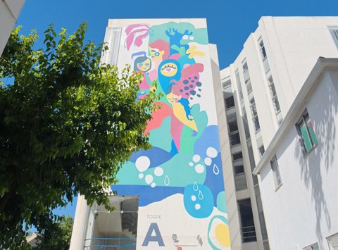 Das Foto zeigt das Gebäude „Torre A“ auf dem Campus der NOVA FCSH in Lissabon. Die Seitenfassade wird von einem großflächigen Wandgemälde geprägt, das sich über mehrere Stockwerke erstreckt. Das Kunstwerk kombiniert menschliche Figuren, Naturmotive und abstrakte Elemente in kräftigen Farben. Ein Baum im linken Bildbereich lockert die Szene auf und verbindet die Architektur mit der umgebenden Natur. Die moderne Hochschulumgebung und das künstlerische Gestaltungselement verleihen dem Campus eine offene, kreative und einladende Atmosphäre.