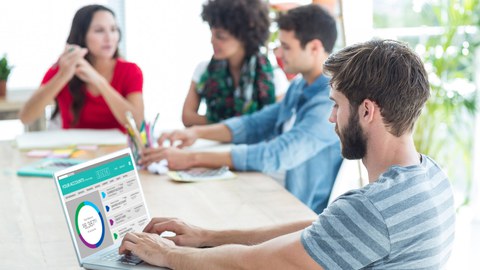 4 junge Menschen am Tisch, einer schaut auf einen Laptop