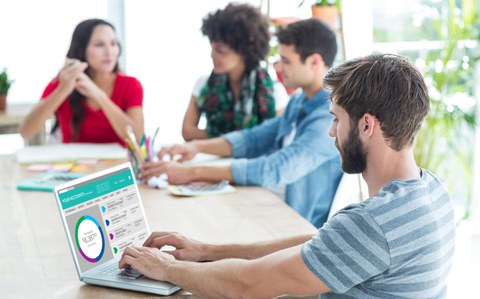 4 junge Menschen am Tisch, einer schaut auf einen Laptop