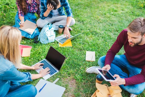 4 Studierende sitzen auf einer Wiese. 3 von Ihnen halten einen Laptop, eine Studentin ein Schreibheft.