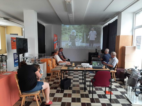 Das Foto zeigt eine Gruppe Menschen, die auf eine Leinwand schauen. Die Leinwand zeigt 2 Personen.