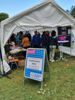 Das Foto zeigt ein großes Festzelt. Im Festzelt sind viele Menschen, die an Tischen stehen oder sich unterhalten. Der Eingang des Festzelts ist offen und davor steht ein Aufsteller mit der Aufschrift Herzlich willkommen auf der Wissenschaftsmeile der TU Dresden und Partnereinrichtungen.