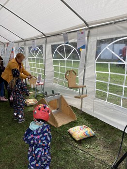 Das Foto zeigt eine Wippe aus Glas und Holz. 2 Kinder stehen davor, eine erwachsene Person hält den einen Sitz der Wippe. Die Wippe steht in einem Festzelt.