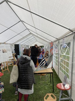 Das Foto zeigt das Innere eines Festzelts. Es stehen einige Menschen im Zelt. Auf einem Tisch steht eine Kugelbahn. Am linken Bildrand stehen Staffeleien mit Plakaten mit Fotos und Text.