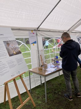 Das Foto zeigt eine Person, die sich Teile einer Ausstellung ansieht. Auf einem Tisch steht ein Glaskasten. Im Glaskasten steht ein Bagger auf einer hellen Fläche. Neben dem Tisch steht eine Staffelei mit einem Plakat. Das Foto wurde in einem Festzelt aufgenommen. 
