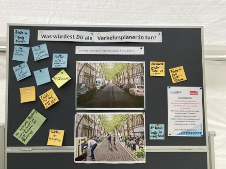 Das Foto zeigt eine Tafel, auf der ganz oben Was würdest DU als Verkehrsplaner:in tun? Darunter steht in kleinerer Schrift Ein Auto benötigt für eine Parkfläche 5 m x 2,50 m. Unter dem Text sind 2 Fotos abgebildet. Das obere Bild zeigte eine Straße mit vielen parkenden Autos. Das untere Bild zeigt die gleiche Straße ohne Autos und mit vielen Menschen. Die Menschen sitzen im Café, gehen spazieren oder beschäftigen sich mit einem Schrank. Neben den beiden Fotos sind Zetteln mit Antworten wie Begrünung, bedachte Straßen, Cafés nur auf einer Seite der Fläche, Rückbau der Versiegelung oder Wasserflächen. Rechts auf der Tafel klebt ein Hinweis auf ein Gewinnspiel.