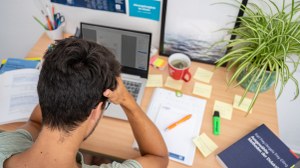 Das Foto zeigt einen Studenten an seinem Schreibtisch. Vor ihm steht sein Laptop und viele weitere Büromaterialien. Mit den Händen stützt er seinen Kopf ab.