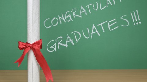 Das Foto zeigt ein Diplom. Dieses steht vor einer Tafel mit der Aufschrift "Congratulations Graduates!!!". Übersetzt bedeutet das: Herzlichen Glückwunsch Absolventinnen und Absolventen.