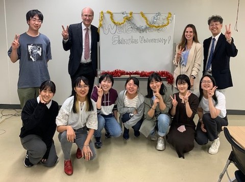 Gruppenbild von japanischen Studenten und deutschen Dozierenden vor einem Schild auf dem "Welcome to Shinshu University" steht.