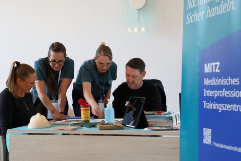 2 Besucher:innen üben chirurgisches Nähen an unterschiedlichen Materialien; unterstützt von 2 Mitarbeiterinnen des MITZ.