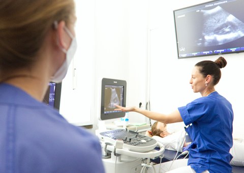 Ärztin führt eine Ultraschalluntersuchung mit dem Sonografiegerät bei einer Person durch, die auf einer Liege liegt; eine weitere Ärztin schaut zu