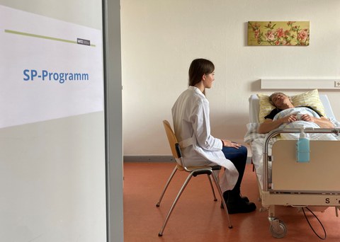 Medizinstudentin im weißen Kittel spricht am Krankenbett mit Simulatonspatient; SP leicht aufgerichtet, Blick zur Studentin; Tür links mit Schild „SP-Programm“.