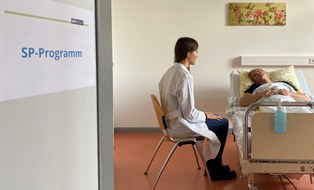 Medizinstudentin im weißen Kittel spricht am Krankenbett mit Simulatonspatient; SP leicht aufgerichtet, Blick zur Studentin; Tür links mit Schild „SP-Programm“.