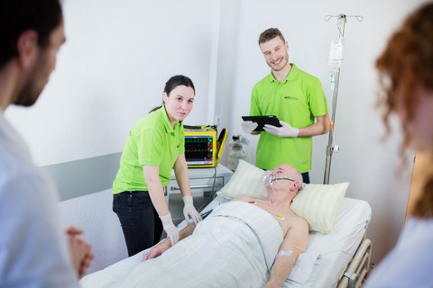 Simulationsperson im Krankenbett und um ihn herum studentische Tutoren und Studierende während einer Lehreinheit