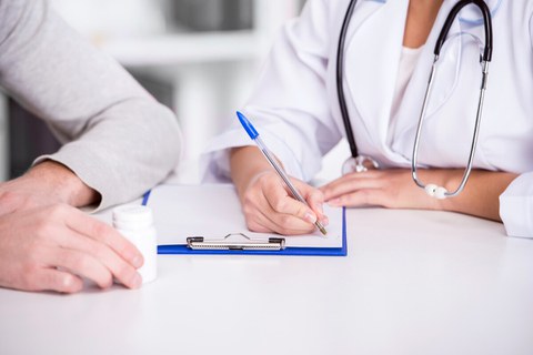 Bild von einer Ärztin, die neben ihrem Patienten sitzt. Dieser hält ein Medikamentenbehälter und sie macht sich Notizen.