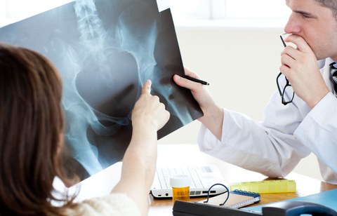Das Foto zeigt einen Arzt, der das Röntgenbild einer Hüfte in der Hand hält. Ihm gegenüber sitzt eine Frau, die mit ihrem Finger auf eine Stelle auf dem Bild zeigt.