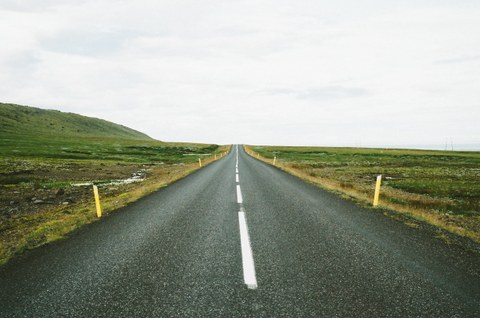 Das Foto zeigt eine lange gerade Straße. Sie verläuft durch eine flache grüne Landschaft.