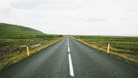 Das Foto zeigt eine lange gerade Straße. Sie verläuft durch eine flache grüne Landschaft.