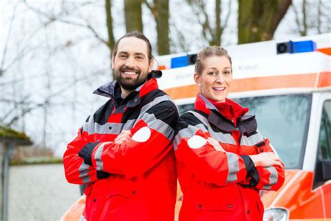 Im Vordergrund sind ein Mann und eine Frau in Uniformen des Rettungsdienstes zu sehen. Im Hintergund ist der Rettungswagen verschwommen erkennbar.