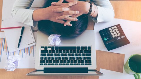 Foto einer Person am Schreibtisch, deren Kopf auf einem aufgeklappten Laptop liegt. Die Person hat die Hände hinter dem Kopf verschränkt und neben ihr liegen Arbeitsutensilien sowie zusammengeknülltes Papier.