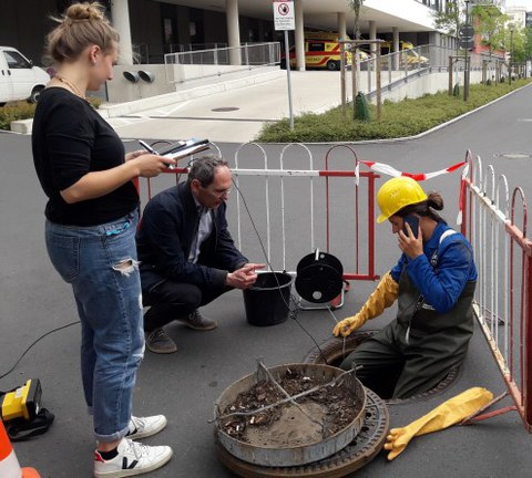 Foto eines geöffneten Gullydeckels auf einem abgesperrten Stück Straße. Eine Person in Schutzkleidung und mit Helm sitzt am Rand des Gullys, die Beine hängen hinein. Eine weitere steht gegenüber und macht auf einem Klemmbrett Notizen. Dahinter hockt eine weitere person und schaut un den Gully. Neben dieser stehen ein Eimer und eine Kabelrolle..