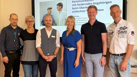 6 Personen (  (MITZ-Dozierende, Schauspielpersonen, Experten Klinik Bavaria Kreischa) im Seminarraum vor einem Smartboard mit Präsentationsfolie "Kommunikation mit fordernden Angehörigen beim Überbringen schlechter Nachrichten"
