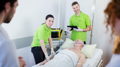 Simulationsperson im Krankenbett und um ihn herum studentische Tutoren und Studierende während einer Lehreinheit