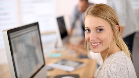 Das Foto zeigt eine junge Frau vor ihrem Computer. Sie hat sich gerade zur Seite gedreht und lächelt in die Kamera.