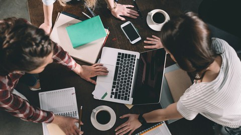 Das Foto zeigt drei junge Frauen an einem Tisch aus der Vogelperspektive. Auf dem Tisch liegen Hefte, Stifte und ein Laptop.