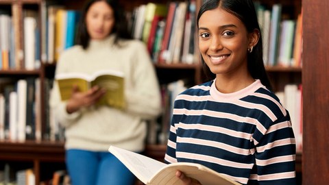 Foto von zwei Studentinnen in der Bibliothek. Die eine im Hintergrund liest und die im Vordergrund lächelt in die Kamera. 