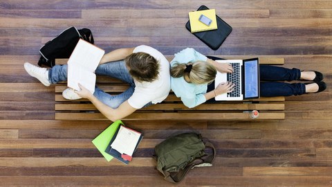 Das Foto zeigt zwei Studierende aus der Vogelperspektive. Sie sitzen Rücken an Rücken auf einer Bank und arbeiten. Um die Bank herum liegen Taschen und weitere Arbeitsmaterialien verteilt.