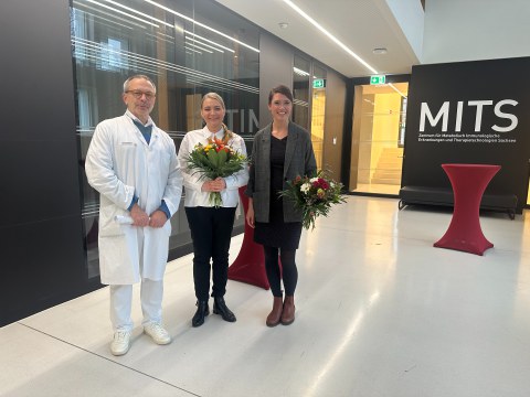 Das Foto zeigt drei Personen, zwei halten einen Blumenstrauß in der Hand.