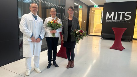 Das Foto zeigt drei Personen, zwei halten einen Blumenstrauß in der Hand.