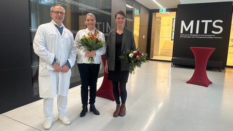 Das Foto zeigt drei Personen, zwei halten einen Blumenstrauß in der Hand.