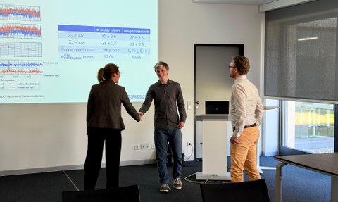 Das Foto zeigt drei Personen. Eine weibliche Person gibt einer männlichen Person die Hand.