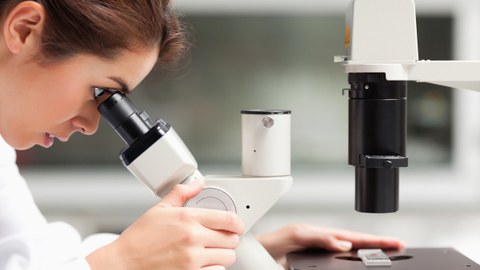 Das Foto zeigt eine junge Frau. Sie schaut gerade durch ein Mikroskop. Auf dem Mikroskop liegt ein Objektträger mit einer roten Flüssigkeit.