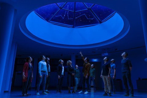 Eine Gruppe von Menschen steht in einem dunklen Raum unter einem runden Dachfenster; durch das Fensterglas ist ein Sternbild sichtbar, das an die Pulslinie erinnert. 