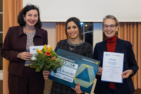 Drei Personen schauen lächelnd in die Kamera; die Personen on der Mitte hält einen Blumenstrauß und einen Spendenscheck mit dem Namen Haidy Hassan darauf; die Person rechts hält eine Urkunde.