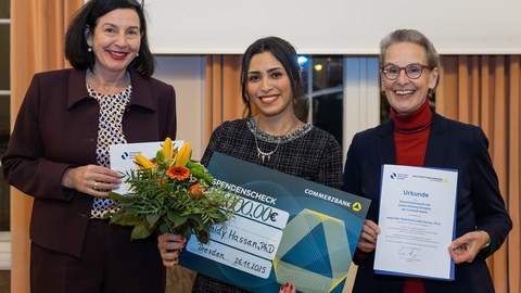 Drei Personen schauen lächelnd in die Kamera; die Personen on der Mitte hält einen Blumenstrauß und einen Spendenscheck mit dem Namen Haidy Hassan darauf; die Person rechts hält eine Urkunde.