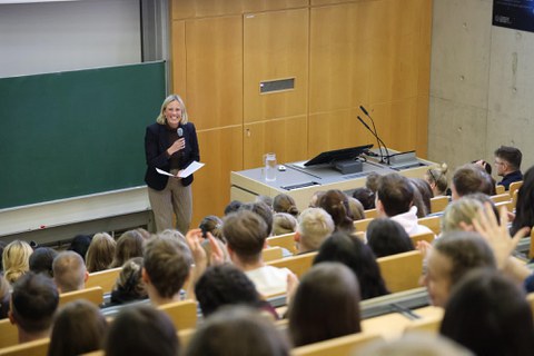 Im Fokus eine lächelnde Person, die vor den Zuhörern in einem Hörsaal spricht.