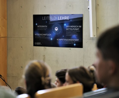 Im Vordergrund unscharfe Silhouetten der Zuhörer, im Hintergrund im Fokus eine Tafel mit dem Titel: Leitbild Lehre
